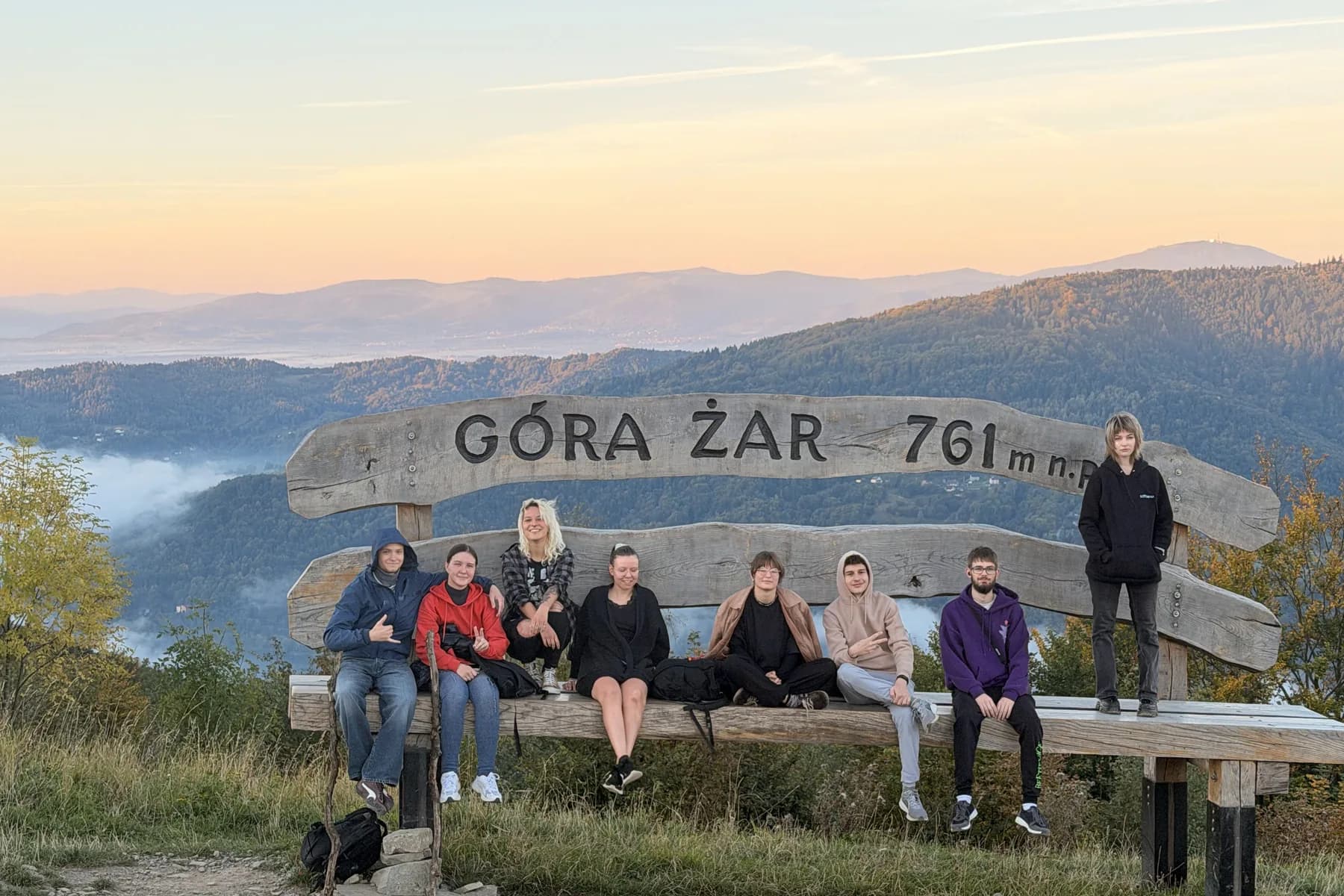 Uczestnicy na ławce pod napisem Góra Żar 761 m n.p.m., o wschodzie słońca